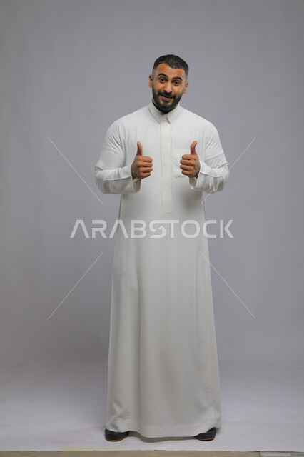 إيماءات الإعجاب والرضى, بورتريه لشاب عربي سعودي خليجي مبتسم يرتدي الثوب التقليدي ويرفع إصبع الابهام نحو الأعلى بعلامة الجودة، الشعور بالموافقه والقبول، الإمتياز والتميز, صورة الجسم بالطول الكامل، خلفية رمادية.