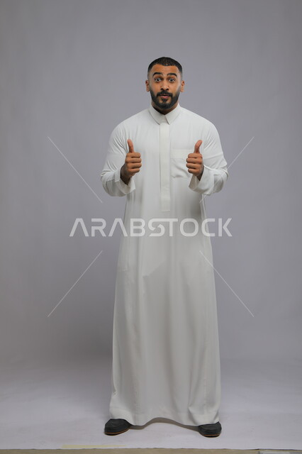 الشعور بالموافقه والقبول، إيماءات الدهشة و الإعجاب والرضى, بورتريه لشاب عربي سعودي خليجي مبتسم يرتدي الثوب التقليدي ويرفع إصبع الابهام نحو الأعلى بعلامة الجودة، الإمتياز والتميز, صورة الجسم بالطول الكامل، خلفية رمادية.