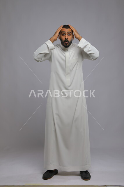 النظر إلى الكاميرا بإيماءات الدهشة والذهول , بورتريه لشاب عربي خليجي سعودي يرتدي الثوب التقليدي يضع يديه على رأسه, النجاح وتحقيق الأهداف, المفاجأة والسعادة بالوصول لأمر ما  , صورة الجسم بالطول الكامل, خلفية رمادية