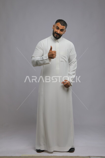 النظر إلى الكاميرا بايماءات القبول والموافقه, بورتريه لرجل عربي خليجي سعودي مبتسم يرتدي الثوب التقليدي ويرفع أصبع  الابهام بعلامة الجودة ،الشعور بالإعجاب والرضا والسعاده، صورة الجسم بالطول الكامل، خلفية رمادية .