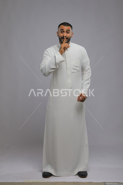 التوقف عن الكلام و الأمر بالصمت و الاستماع، بورتريه لرجل عربي سعودي خليجي يرتدي الثوب التقليدي ينظر إلى الكاميرا ويرفع اصبعه الى فمه, إيماءات يدين تدل على طلب الهدوء و عدم الإزعاج, صورة الجسم بالطول الكامل، خلفية رمادية.