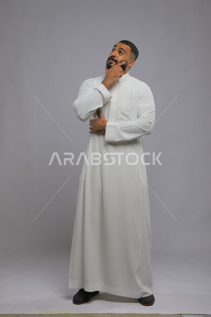 التفكير و التأمل و شرود الذهن والتخطيط للمستقبل, بورتريه لشاب عربي خليجي سعودي مبتسم يرتدي الثوب التقليدي يقوم بإيجاد حل لمشكلة ما, إيماءات التمعن والتركيز، صورة الجسم بالطول الكامل،  خلفية رمادية .