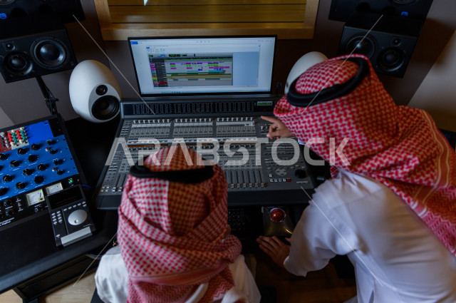 شابان سعوديان في غرفة خارج الاستديو ،يتم معالجةالاصوات المسجلة داخل الاستديو عن طريق برمجيات صوتية من خلال جهاز الحاسوب المخصص ، استديو تسجيل صوتي ، بث مباشر ، فريق الاعداد ، الاذاعة الصوتية ، نشرات اخبارية