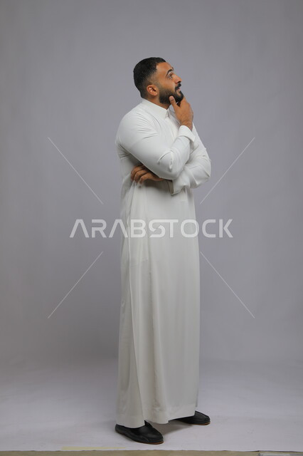 التأمل التفكير و شرود الذهن والتخطيط للمستقبل, بورتريه لشاب عربي خليجي سعودي يرتدي الثوب التقليدي يقوم بإيجاد حل لمشكلة ما, إيماءات التمعن والتركيز، صورة الجسم بالطول الكامل،  خلفية رمادية .