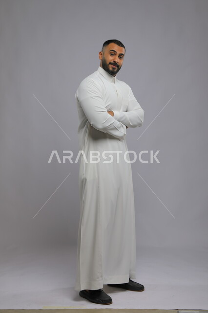 الوقوف بحركات أيدي متقاطعه ، مفهوم الرجولة والأناقه، بورتريه لشاب وسيم عربي خليجي سعودي مبتسم يرتدي الثوب التقليدي ينظر إلى الكاميرا بإيماءات السرور و الثقه بالنفس، الاهتمام بالمظهر الخارجي، صورة الجسم بالطول الكامل، خلفية رمادية.
