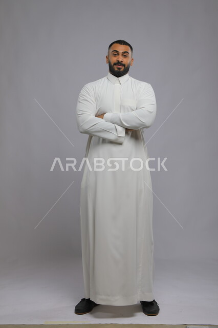 مفهوم الرجولة والأناقه، الوقوف بحركات أيدي متقاطعه ، بورتريه لشاب وسيم عربي خليجي سعودي مبتسم يرتدي الثوب التقليدي ينظر إلى الكاميرا بإيماءات السرور و الثقه بالنفس، الاهتمام بالمظهر الخارجي، صورة الجسم بالطول الكامل، خلفية رمادية.
