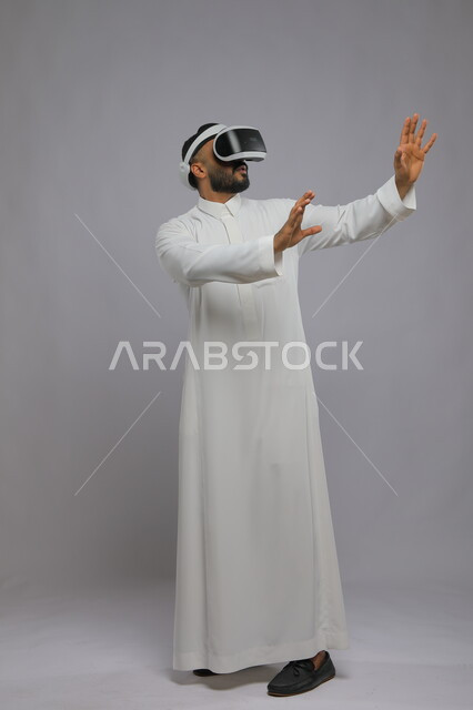 Interaction and simulation of the imaginary world, portrait of a Saudi Gulf Arab young man wearing a traditional dress, watching movies through 3D virtual reality glasses, enthusiasm, integration, and trying to catch something, full-length body image, gray background