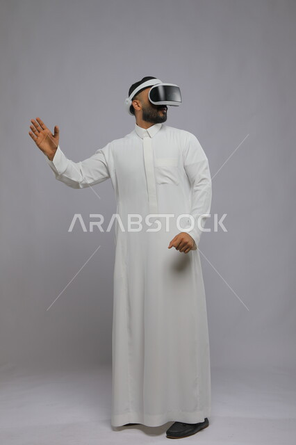 مشاهدة الأفلام عبر نظارة الواقع الافتراضي, التفاعل والاندماج ومحاكاة العالم الخيالي, بورتريه لرجل عربي خليجي سعودي يرتدي الثوب التقليدي والنظارة ثلاثية الابعاد يرفع يديه للأعلى, صورة الجسم بالكامل, خلفية رمادية
