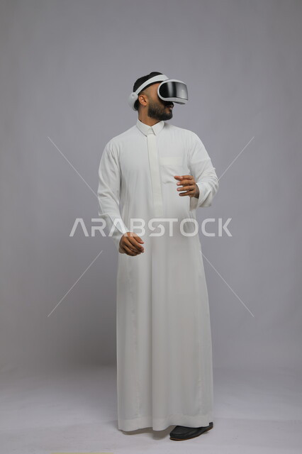 التفاعل والاندماج ومحاكاة العالم الخيالي, بورتريه لرجل عربي خليجي سعودي يرتدي الثوب التقليدي والنظارة ثلاثية الابعاد يرفع يديه للأعلى, مشاهدة الأفلام عبر نظارة الواقع الافتراضي, صورة الجسم بالكامل, خلفية رمادية