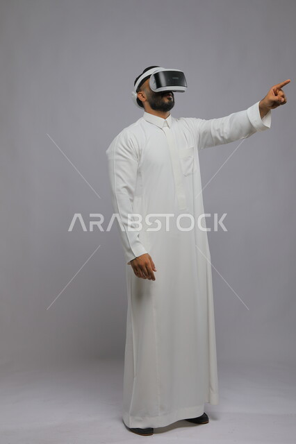 محاكاة العالم الخيالي, استخدام حاسة اللمس لملامسة عناصر هولوجرامية بإيماءات النقر, بورتريه لرجل عربي خليجي سعودي يرتدي الثوب التقليدي ونظارة الواقع الافتراضي ثلاثية الأبعاد يشير إلى شيء ما بأصبعه السبابة, صورة الطول الكامل للجسم, خلفية رمادية