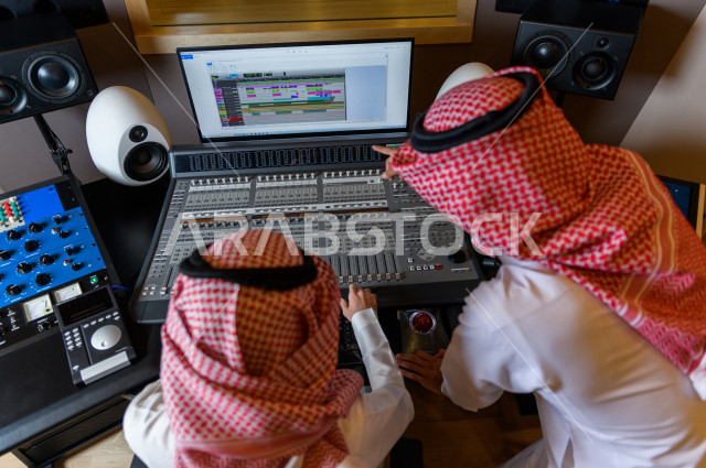 شابان سعوديان في غرفة خارج الاستديو ،يتم معالجةالاصوات المسجلة داخل الاستديو عن طريق برمجيات صوتية من خلال جهاز الحاسوب المخصص ، استديو تسجيل صوتي ، بث مباشر ، فريق الاعداد ، الاذاعة الصوتية ، نشرات اخبارية