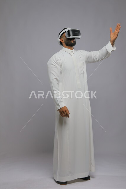 المتعه والتسلية بمشاهدة الافلام ومحاكاة العالم الخيالي, بورتريه لشاب عربي خليجي سعودي يرتدي الثوب التقليدي يقوم بمشاهدة أفلام عبر نظارة الواقع الافتراضي ثلاثية الابعاد, محاولة لمس شيء ما بإيماءات رفع الكف, صورة الجسم بالطول الكامل, خلفية رمادية