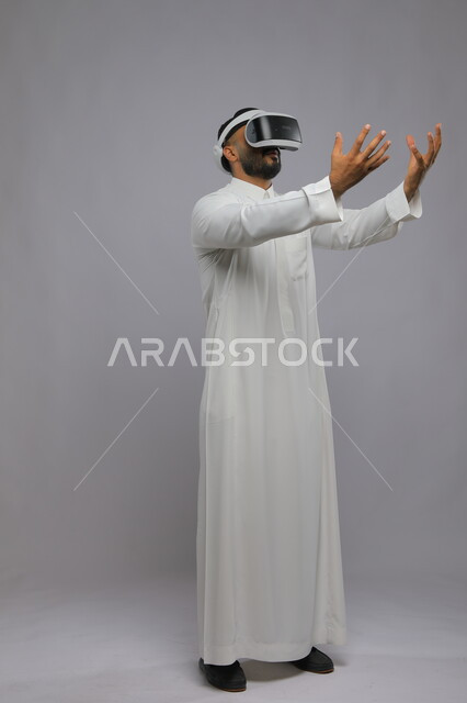 الاندماج والحماس ومحاولة امساك شيء ما, التفاعل ومحاكاة العالم الخيالي, بورتريه لشاب عربي خليجي سعودي يرتدي الثوب التقليدي يقوم بمشاهدة أفلام عبر نظارة الواقع الافتراضي ثلاثية الأبعاد, صورة الجسم بالطول الكامل, خلفية رمادية