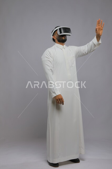 المتعه والتسلية بمشاهدة الافلام ومحاكاة العالم الخيالي, بورتريه لشاب عربي خليجي سعودي يرتدي الثوب التقليدي يقوم بمشاهدة أفلام عبر نظارة الواقع الافتراضي ثلاثية الابعاد, محاولة لمس شيء ما بإيماءات رفع الكف, صورة الجسم بالطول الكامل, خلفية رمادية