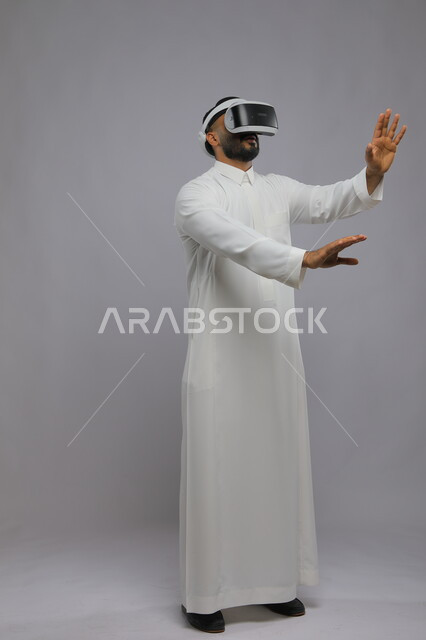 الحماس والاندماج ومحاولة امساك شيء ما, التفاعل ومحاكاة العالم الخيالي, بورتريه لشاب عربي خليجي سعودي يرتدي الثوب التقليدي يقوم بمشاهدة أفلام عبر نظارة الواقع الافتراضي ثلاثية الأبعاد, صورة الجسم بالطول الكامل, خلفية رمادية