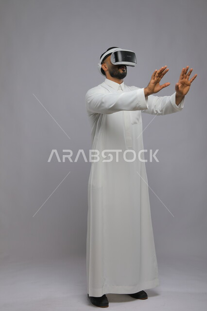 Enthusiasm, integration, and trying to catch something, interaction and simulation of the imaginary world, portrait of a Saudi Gulf Arab young man wearing a traditional dress, watching movies through 3D virtual reality glasses, full-length body image, gray background