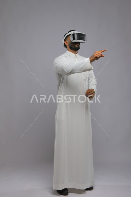 استخدام حاسة اللمس لملامسة عناصر هولوجرامية بإيماءات النقر, بورتريه لرجل عربي خليجي سعودي يرتدي الثوب التقليدي ونظارة الواقع الافتراضي ثلاثية الأبعاد يشير إلى شيء ما بأصبعه السبابة, محاكاة العالم الخيالي, صورة الطول الكامل للجسم, خلفية رمادية