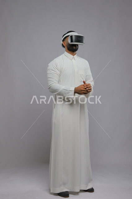 الاندماج والتفاعل ومحاكاة العالم الخيالي, بورتريه لشاب عربي خليجي سعودي يرتدي الثوب التقليدي يقوم بمشاهدة أفلام عبر نظارة الواقع الافتراضي ثلاثية الأبعاد, صورة الجسم بالطول الكامل, خلفية رمادية