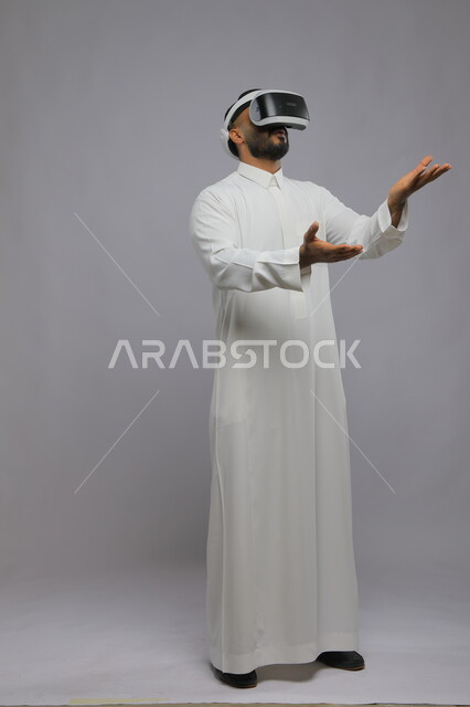 التفاعل ومحاكاة العالم الخيالي, بورتريه لشاب عربي خليجي سعودي يرتدي الثوب التقليدي يقوم بمشاهدة أفلام عبر نظارة الواقع الافتراضي ثلاثية الأبعاد, الحماس والاندماج ومحاولة امساك شيء ما, صورة الجسم بالطول الكامل, خلفية رمادية