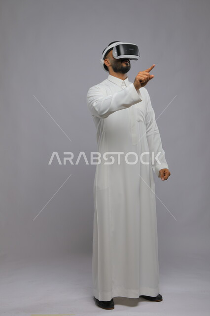 استخدام حاسة اللمس لملامسة عناصر هولوجرامية بإيماءات النقر, بورتريه لرجل عربي خليجي سعودي يرتدي الثوب التقليدي ونظارة الواقع الافتراضي ثلاثية الأبعاد يشير إلى شيء ما بأصبعه السبابة, محاكاة العالم الخيالي, صورة الجسم بالطول الكامل, خلفية رمادية