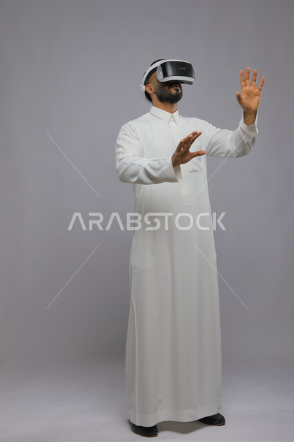 التفاعل ومحاكاة العالم الخيالي, بورتريه لشاب عربي خليجي سعودي يرتدي الثوب التقليدي يقوم بمشاهدة أفلام عبر نظارة الواقع الافتراضي ثلاثية الأبعاد, الحماس والاندماج ومحاولة امساك شيء ما, صورة الجسم بالطول الكامل, خلفية رمادية
