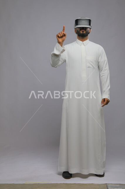 الاندماج ومحاكاة العالم الخيالي, استخدام حاسة اللمس لملامسة عناصر هولوجرامية, بورتريه لرجل عربي خليجي سعودي يرتدي الثوب التقليدي ونظارة الواقع الافتراضي ثلاثية الأبعاد يشير إلى شيء ما بأصبعه السبابة, صورة الجسم بالكامل, خلفية رمادية
