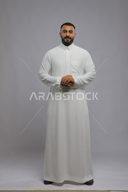 الاهتمام بالمظهر الخارجي، الوقوف بحركات أيدي متماسكة ، مفهوم الرجولة والأناقه، بورتريه لشاب وسيم عربي خليجي سعودي مبتسم يرتدي الثوب التقليدي ينظر إلى الكاميرا بإيماءات السرور و الثقه بالنفس، صورة الجسم بالطول الكامل، خلفية رمادية.