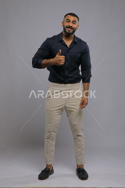الشعور بالرضا والاعجاب والسعاده، النظر إلى الكاميرا بايماءات القبول والموافقه, بورتريه لشاب وسيم عربي خليجي سعودي مبتسم يرتدي الزي الكاجوال ويرفع أصبع الابهام بعلامة الجودة ،صورة الجسم بالطول الكامل، خلفية رمادية .