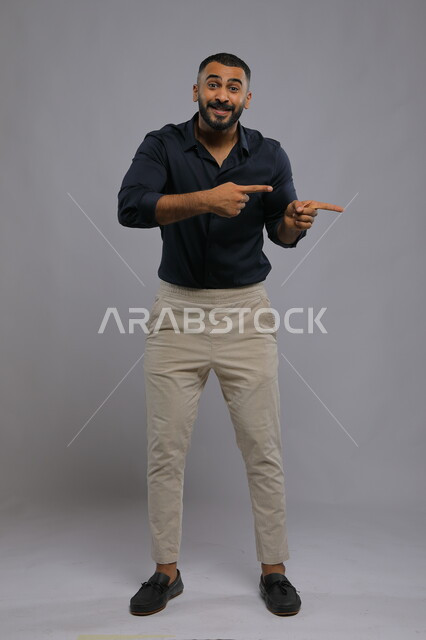 Expressions of confirmation and locating a place, pointing to something with gestures of pleasure, portrait of a smiling Saudi Gulf Arab man wearing a casual outfit, raising his index finger to choose and looking at the camera, full body portrait, gray background