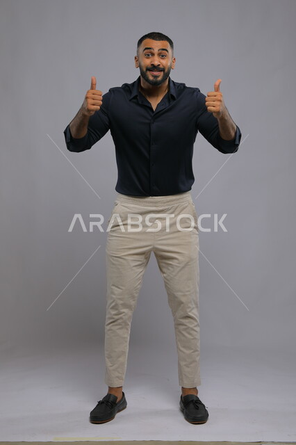 النظر إلى الكاميرا بايماءات القبول والموافقه, الشعور بالإعجاب والرضا والسعاده، بورتريه لشاب وسيم عربي خليجي سعودي مبتسم يرتدي الزي الكاجوال ويرفع أصبع الابهام بعلامة الجودة ،صورة الجسم بالطول الكامل، خلفية رمادية .
