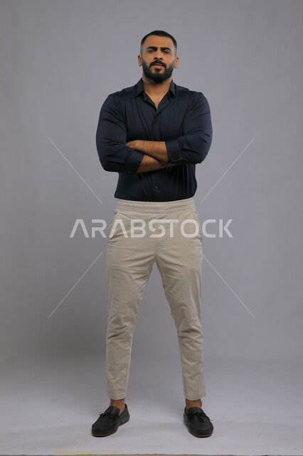 Expressions of frowning, dissatisfaction and anger, standing upright and self-confident, portrait of a handsome Saudi Gulf Arab young man wearing casual clothes and standing upright, looking at the camera with crossed hand gestures, full-length body portrait, gray background