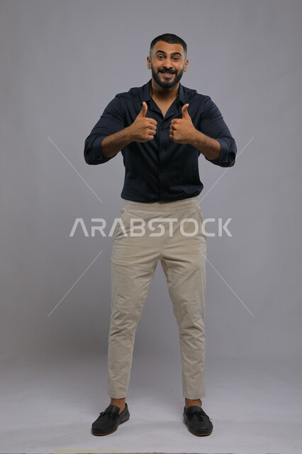 Feeling of admiration, satisfaction, and happiness, looking at the camera with gestures of acceptance and approval, portrait of a handsome, Saudi Gulf Arab young man wearing a casual outfit and raising a thumb with a quality sign, full-length body photo, gray background.