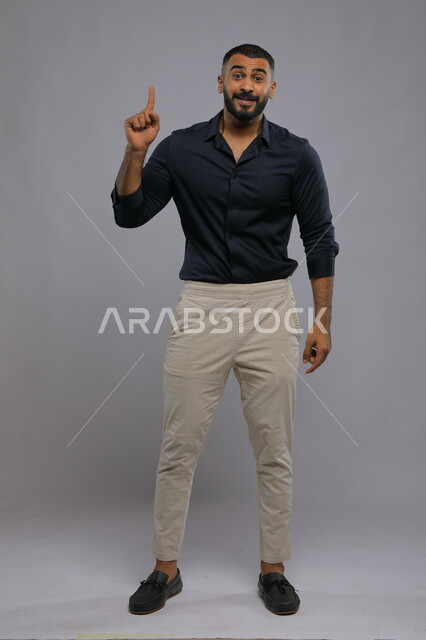  ايماءات الاستفسار والاشارة إلى شيء ما, الوقوف باستقامة والنظر إلى الكاميرا, بورتريه لرجل عربي خليجي سعودي مبتسم يرتدي الزي الكاجوال يرفع اصبعه للأعلى بإيماءات التوضيح, صورة الجسم بالطول الكامل , خلفية رمادية
