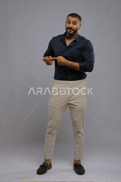 الاشارة إلى شيء ما بإيماءات السرور ,تعابير التأكد والتحديد,  بورتريه لرجل عربي خليجي سعودي مبتسم يرتدي الزي الكاجوال يرفع إصبعه السبابة للاختيار وينظر إلى الكاميرا, صورة للجسم بالكامل, خلفية رمادية