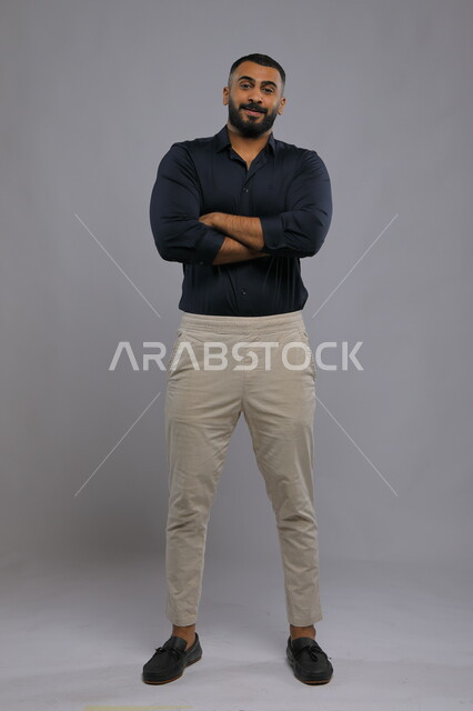 Standing upright and self-confident, a portrait of a handsome Saudi Gulf Arab young man wearing casual clothes and standing upright, looking at the camera with crossed hand gestures, the concept of elegance and concern for external appearance, full-length body portrait, gray background