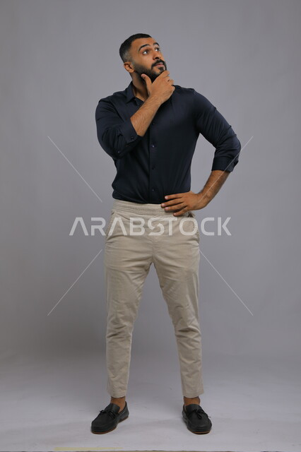 Gestures of thinking about something, contemplating, contemplating and planning for the future, portrait of a handsome Saudi Gulf Arab young man wearing casual clothes, placing his hand on his waist and looking up with an expression of contemplation and concentration to find a solution to a problem, full-body portrait, gray background