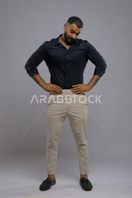 ايماءات العبوس والانزعاج, بورتريه لشاب وسيم عربي خليجي سعودي يرتدي الزي الكاجوال يضع يديه على خصره و ينظر إلى الأسفل ،الوقوف بتعابير تدل على الانتظار، الملل والشرود والتفكير، صورة الجسم بالطول الكامل، خلفية رمادية