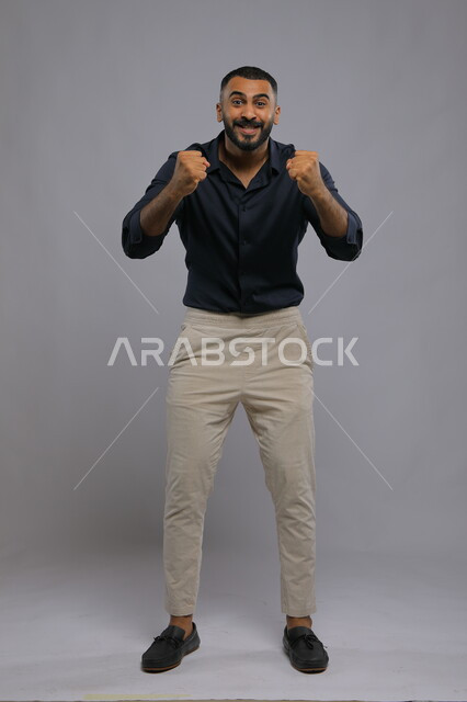 حركات تدل على الكسب والربح وانجاز المهام، بورتريه لشاب عربي خليجي سعودي يرتدي الزي الكاجوال و يرفع يديه للأعلى بتعابير النجاح و تحقيق الأهداف ,النظر إلى الكاميرا بإيماءات الفرح والانتصار, صورة الجسم بالطول الكامل, خلفية رمادية