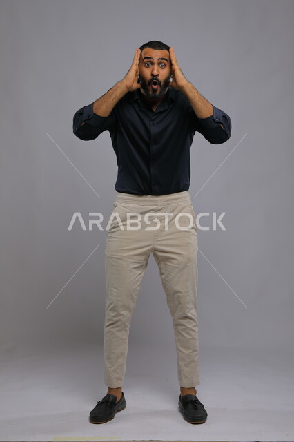 التفاجؤ من شيء ما، الشعور بالدهشة والتعجب والصدمة والذهول , بورتريه لشاب عربي خليجي سعودي يرتدي الزي الكاجوال و يرفع يديه على رأسه بتعابير النجاح وتحقيق الأهداف, النظر إلى الكاميرا بإيماءات الفرح والسرور , صورة الجسم بالطول الكامل, خلفية رمادية