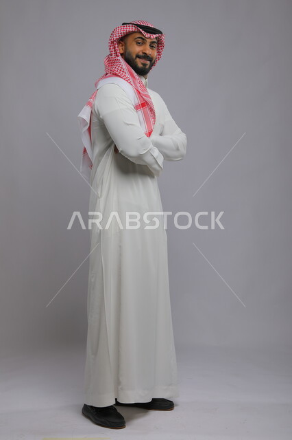 الوقوف بحركات أيدي متقاطعة، مفهوم الأناقة والرجولة، بورتريه لرجل عربي خليجي سعودي مبتسم يرتدي الثوب والشماغ التقليدي ينظر إلى الكاميرا بإيماءات الثقه بالنفس، الاهتمام بالمظهر الخارجي، صورة لكامل الجسم، خلفية رمادية.