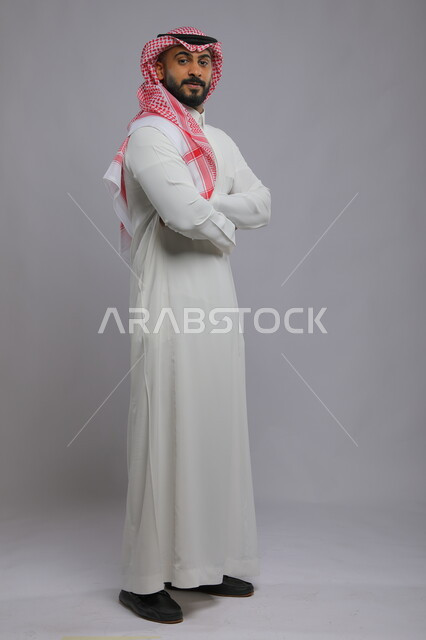 الاهتمام بالمظهر الخارجي، الوقوف بحركات أيدي متقاطعة، مفهوم الأناقة والرجولة، بورتريه لرجل عربي خليجي سعودي مبتسم يرتدي الثوب والشماغ التقليدي ينظر إلى الكاميرا بإيماءات الثقه بالنفس، صورة لكامل الجسم، خلفية رمادية.