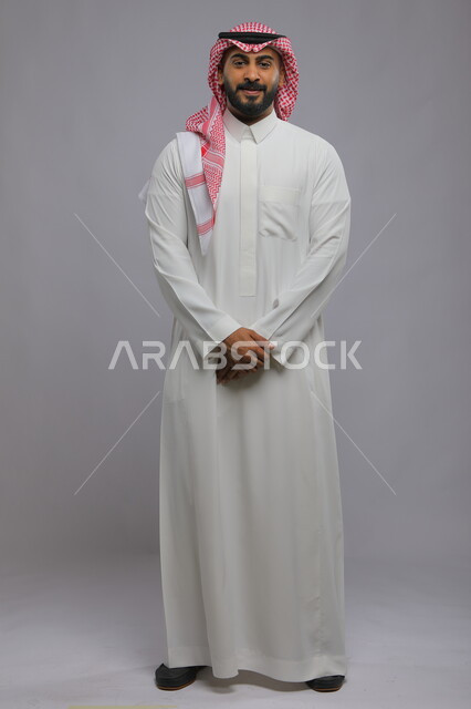 الوقوف باستقامة بحركات أيدي متماسكة، الاهتمام بالمظهر الخارجي، مفهوم الرجولة والأناقه، بورتريه لرجل عربي خليجي سعودي مبتسم يرتدي الزي التقليدي ينظر إلى الكاميرا بإيماءات الثقه بالنفس، صورة الجسم بالطول الكامل، خلفية رمادية.