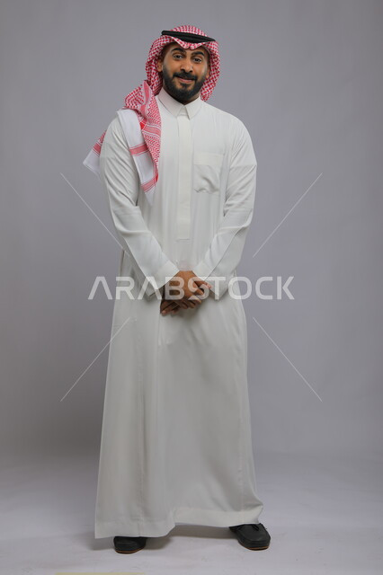 مفهوم الرجولة والأناقه، الوقوف باستقامة بحركات أيدي متماسكة، الاهتمام بالمظهر الخارجي، بورتريه لرجل عربي خليجي سعودي مبتسم يرتدي الزي التقليدي ينظر إلى الكاميرا بإيماءات السرور والدهشة، صورة الجسم بالطول الكامل، خلفية رمادية.