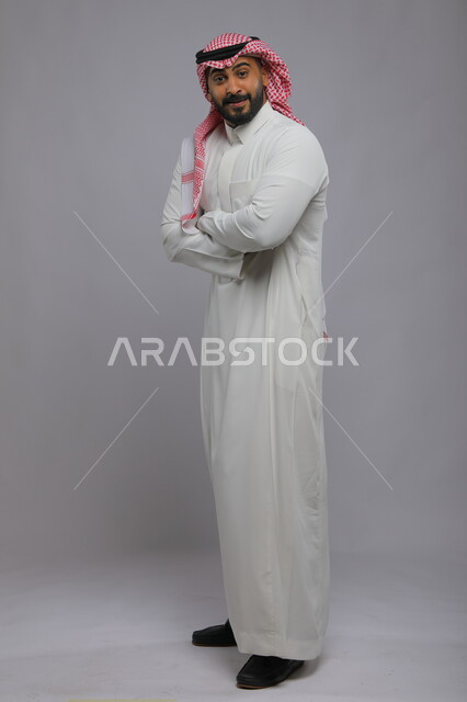 الوقوف بحركات أيدي متقاطعة، مفهوم الأناقة والرجولة، بورتريه لرجل عربي خليجي سعودي يرتدي الثوب والشماغ التقليدي ينظر إلى الكاميرا بإيماءات تدل على الدهشة والتعجب والانتظار، الاهتمام بالمظهر الخارجي، صورة لكامل الجسم، خلفية رمادية.