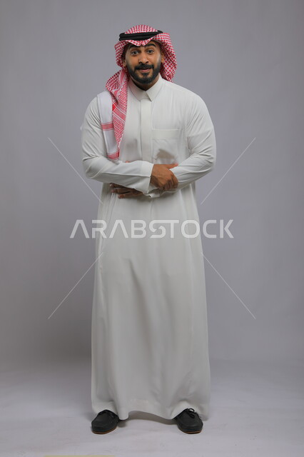 إيماءات وجه تدل على الدهشة والتعجب والذهول، مفهوم الأناقة والرجولة، بورتريه لرجل عربي خليجي سعودي يرتدي الثوب والشماغ التقليدي ينظر إلى الكاميرا يقف بحركات أيدي متماسكة، الاهتمام بالمظهر الخارجي، صورة لكامل الجسم، خلفية رمادية.