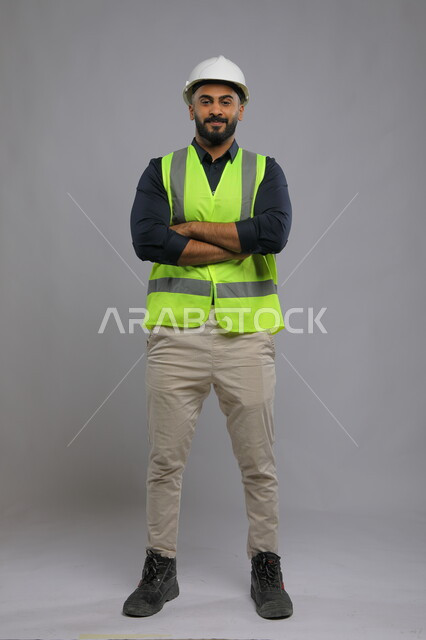 التقدم والتطور في المجال الهندسي السعودي، بورتريه لمهندس عربي سعودي خليجي يرتدي الخوذة وسترة الحماية متكتف اليدين ينظر الى الكاميرا بابتسامة، تصميم وتنفيذ المشاريع الهندسية الكبرى، صورة الجسم بالطول الكامل، خلفية رمادي