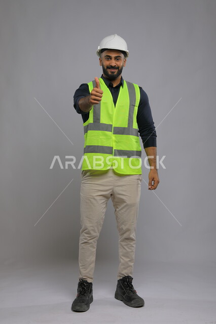 ايماءات الموافقة والقبول، بورتريه لمهندس عربي سعودي خليجي يرتدي الخوذة وسترة الحماية يصنع بيده اشارة الاعجاب، الوظائف الهندسية في الشركات السعودية، صورة الجسم بالطول الكامل، خلفية رمادية
