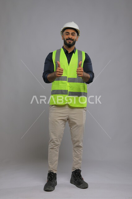 ايماءات الموافقة والقبول، بورتريه لمهندس عربي سعودي خليجي يرتدي الخوذة وسترة الحماية  يصنع بيديه اشارة الاعجاب، الوظائف الهندسية في الشركات السعودية، صورة الجسم بالطول الكامل، خلفية رمادية