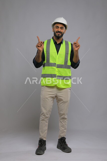 The field of construction and engineering projects,, Portrait of a Saudi Gulf Arab engineer wearing a helmet and protection vest, raising his index finger up, engineering jobs in Saudi companies, full-length body portrait, gray background