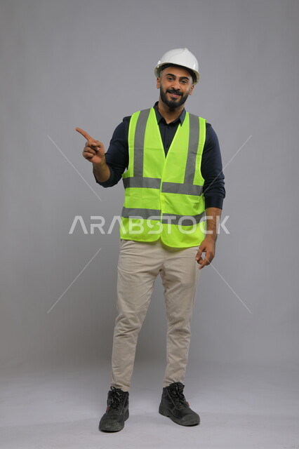 Progress and development in the Saudi engineering field, a portrait of a Saudi Gulf Arab engineer wearing a helmet and a protective vest pointing his finger somewhere, the importance of engineering in developing and improving the infrastructure of cities, full-length body image, gray background
